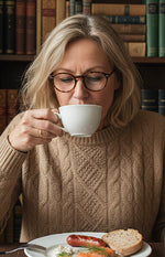 A woman with glasses drinks from a white cup, with a plate of food in front of her.