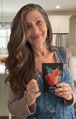 A smiling woman with long hair wearing overalls holds up a pouch of Pipi Tea in a kitchen.